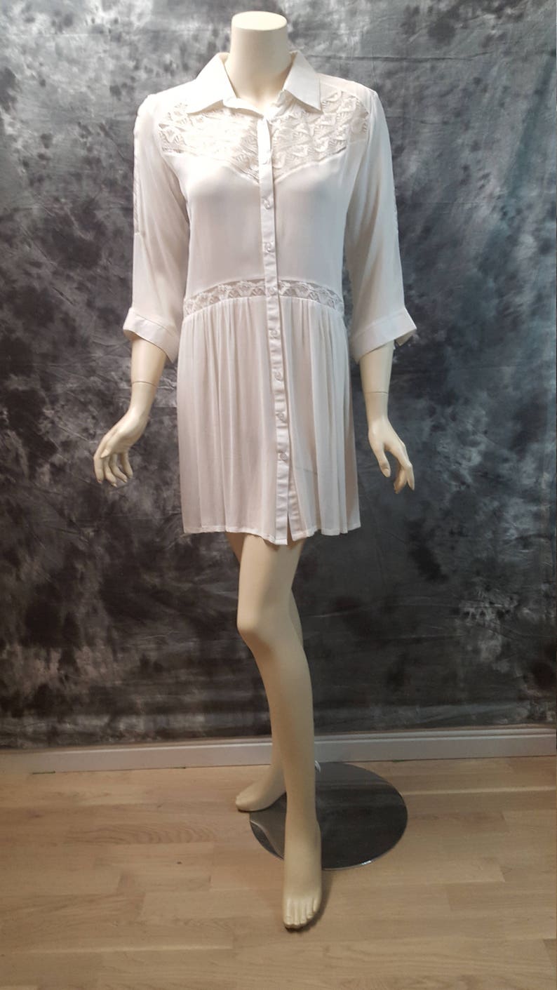 May include: A white button-down shirt dress with lace detailing on the bodice and sleeves. The dress has a relaxed fit and a slightly flared skirt.
