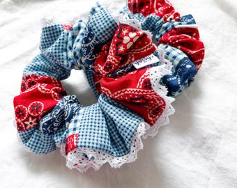 Puede incluir: Un coletero de tela con un diseño de patchwork que combina patrones de bandana en rojo y azul con cuadros vichy en azul claro. El accesorio está adornado con un delicado borde de encaje blanco e incluye una pequeña etiqueta blanca.