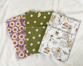 Puede incluir: Tres bolsas de tela con cierres de botón. Una es de color malva con margaritas blancas, otra es verde oliva con margaritas blancas y la tercera es blanca con un estampado botánico vintage. Las bolsas son probablemente para llevar objetos pequeños.