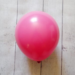 May include: A bright pink balloon, fully inflated, is centered in the frame. The balloon has a smooth, glossy surface and a rounded shape. The background is a light-colored wooden surface with vertical planks.