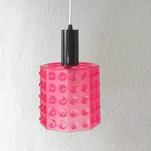 Könnte beinhalten: Eine rosa Pendelleuchte mit einem sechseckigen Schirm, der ein strukturiertes Muster aus kleinen Kreisen aufweist. Die Leuchte ist an einem weißen Kabel und einer schwarzen Deckenhalterung befestigt.