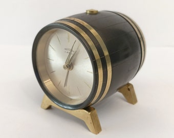 Könnte beinhalten: Eine kleine, schwarze und goldene Uhr in Form eines Fasses. Das Zifferblatt der Uhr ist weiß mit schwarzen Zeigern und dem Text "Westclox" in Schwarz.