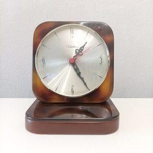 May include: A square clock with a brown and white tortoise shell design. The clock face is silver with black hands and the brand name "Diehl" is printed on the face.