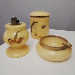Könnte beinhalten: Ein Set aus drei Rauchzubehör aus Alabaster. Das Set enthält ein Feuerzeug, einen Aschenbecher und einen Zigarettenhalter. Das Feuerzeug ist mit einer Pfeife und einer Zigarette verziert. Der Aschenbecher ist rund und hat einen goldenen Rand. Der Zigarettenhalter ist zylindrisch und hat einen goldenen Deckel.