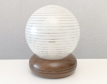 Puede incluir: Una lámpara de globo blanca con rayas horizontales sobre una base de madera marrón.