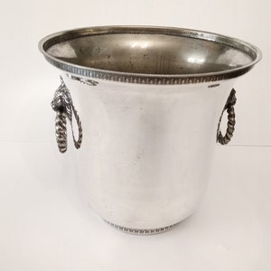 Può includere: Un secchiello per il ghiaccio in argento con due manici in corda con dettagli a testa di leone. Il secchiello ha un bordo perlato.