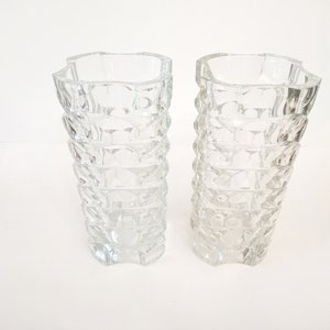 May include: Two clear glass vases with a textured, geometric design. The vases have a square top and a flared, ridged body. The clear glass allows light to pass through, creating a sparkling effect. These vases are suitable for floral arrangements.
