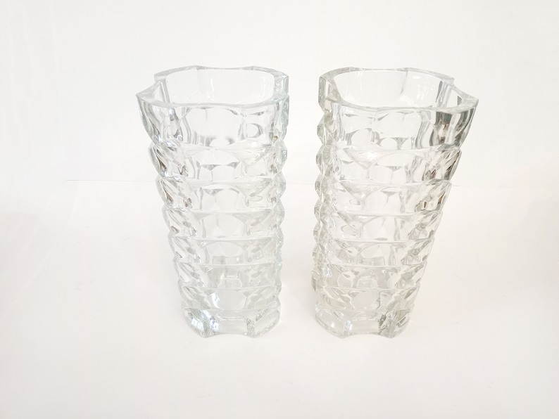 May include: Two clear glass vases with a textured, geometric design. The vases have a square top and a flared, ridged body. The clear glass allows light to pass through, creating a sparkling effect. These vases are suitable for floral arrangements.
