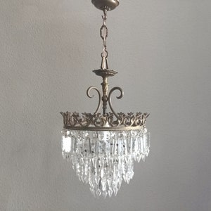 May include: A gold-toned chandelier with a crystal prism design. The chandelier has a decorative crown-like top and a tiered design with multiple rows of crystals.