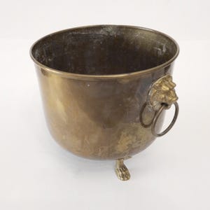 May include: A brass planter with lion head handles and three feet. The planter is round and has a smooth, shiny surface.