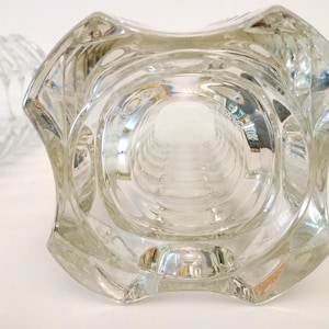 May include: Clear glass vase with a unique, geometric design. The vase has a wide, square-shaped top that tapers down to a cylindrical body. The glass is thick and has a textured surface, creating interesting light reflections. The vase is likely for decorative purposes.