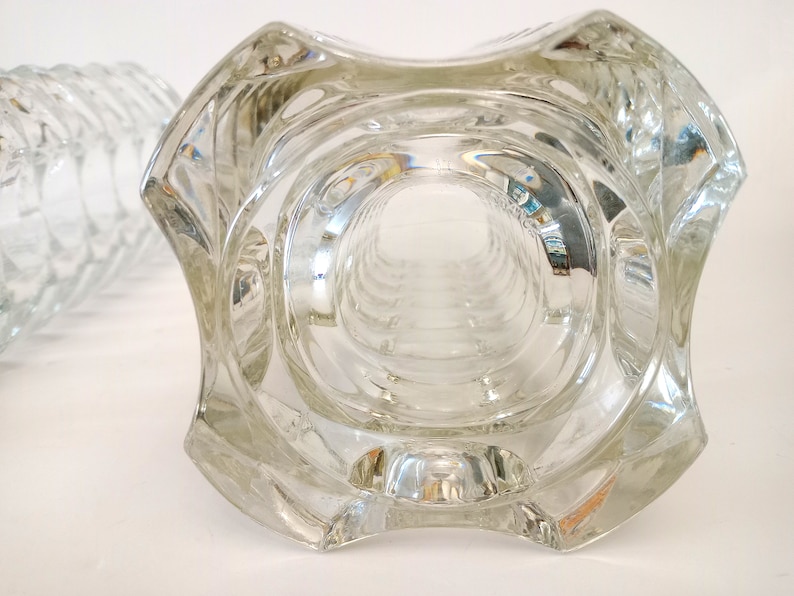 May include: Clear glass vase with a unique, geometric design. The vase has a wide, square-shaped top that tapers down to a cylindrical body. The glass is thick and has a textured surface, creating interesting light reflections. The vase is likely for decorative purposes.