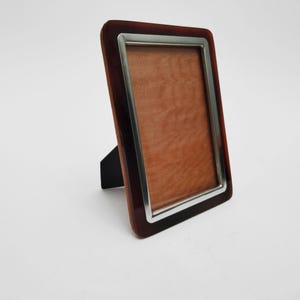 Può includere: Una cornice fotografica rettangolare con un bordo in legno marrone scuro e una finitura in metallo color argento. La cornice ha un retro marrone e un cavalletto nero. La cornice è progettata per contenere una fotografia.