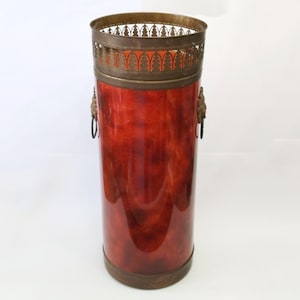 May include: A tall, cylindrical umbrella stand with a rich, reddish-brown finish. It features ornate brass accents, including a decorative rim, lion's head handles, and a base. The stand is approximately 71 cm tall.