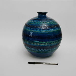 May include: A round, blue ceramic vase with a textured surface and a narrow neck. The vase features horizontal bands of varying shades of blue and teal, with decorative patterns. A black pen rests beside the vase.