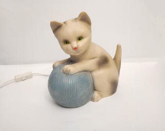 May include: A white ceramic cat figurine with green eyes and a red nose, sitting on a blue ball of yarn. The cat has a tail and is holding the yarn ball with its paws. The figurine is a nightlight with a white cord and a switch.