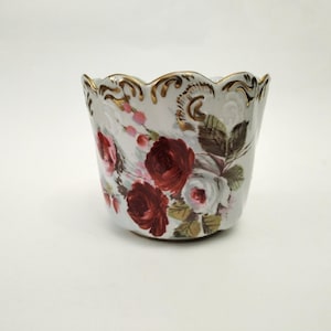 Könnte beinhalten: Ein weißer Keramiktopf mit gewelltem Rand und goldenem Besatz. Der Topf ist mit einem Blumenmuster mit roten und weißen Rosen, grünen Blättern und rosa Akzenten verziert. Das Gesamtdesign ist verziert und vintage.