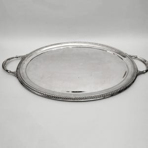Könnte beinhalten: Ovale Serviertablett aus Silber mit zwei Griffen. Das Tablett hat eine reflektierende Oberfläche und einen dekorativen, seilartigen Rand. Die Griffe sind gebogen und an den Seiten befestigt. Das Tablett befindet sich auf weißem Hintergrund.