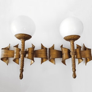May include: Two gold-colored sconces with white globe shades. The sconces have a decorative ribbon design and a textured finish. The sconces are mounted on a white wall.