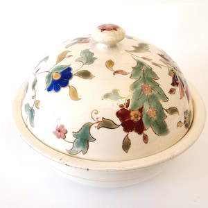 Könnte beinhalten: Eine weiße, überdachte Keramikschale mit einem Blumendesign in Blautönen, Grün, Rot und Gold. Die Schale hat einen gewölbten Deckel und einen runden Boden.