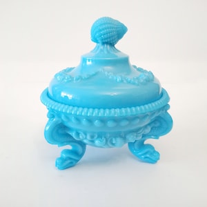 Può includere: Una zuccheriera di vetro blu con un coperchio a forma di conchiglia e tre gambe. La ciotola è decorata con dettagli in rilievo di conchiglie e fiori.