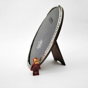 Op de afbeelding: Ovale fotolijst met een zilverkleurige decoratieve rand en een donkerhouten standaard. Het frame staat schuin en toont een donker interieur. Een klein Iron Man Lego-figuur staat voor het frame.