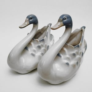 Peut inclure: Deux figurines de cygnes en céramique. Les cygnes ont des corps blancs, des têtes et des becs gris-bleu, et des accents gris-bleu sur leurs ailes. Les cygnes sont placés côte à côte, face à la même direction.