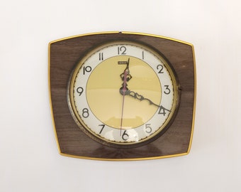 May include: A brown wood framed clock with a gold trim and a white face with black numbers. The clock face has a yellow background and the word "HERCO" printed on it.