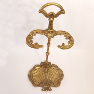 Może przedstawiać: Złoty stojak na parasole z ozdobnym wzorem. Stojak ma okrągłą podstawę z ozdobnym wzorem i pojedynczy pionowy słupek z dwoma zakrzywionymi ramionami.