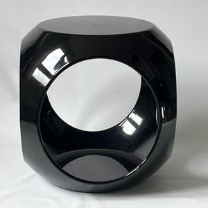 K&ouml;nnte beinhalten: Ein schwarzer, gl&auml;nzender, geometrischer Hocker mit einem runden Ausschnitt in der Mitte. Der Hocker hat ein modernes, minimalistisches Design.