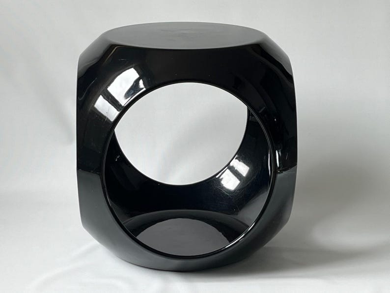 K&ouml;nnte beinhalten: Ein schwarzer, gl&auml;nzender, geometrischer Hocker mit einem runden Ausschnitt in der Mitte. Der Hocker hat ein modernes, minimalistisches Design.