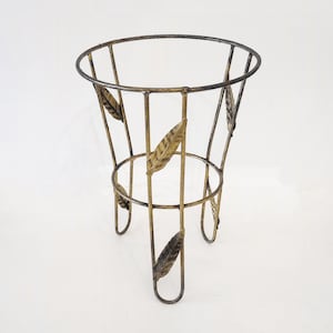 May include: A gold-colored metal plant stand with a round top and three legs. The stand features decorative leaf accents and a cage-like design. The stand is approximately 24 inches tall.