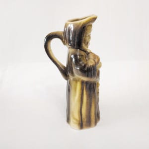 May include: A ceramic pitcher shaped like a monk with a brown and yellow glaze. The pitcher has a handle and a spout.