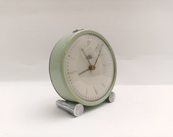 Puede incluir: Un reloj despertador vintage verde con una esfera blanca y marcadores de hora romanos negros. El reloj tiene un logotipo de sol plateado y una aguja de segundos plateada.