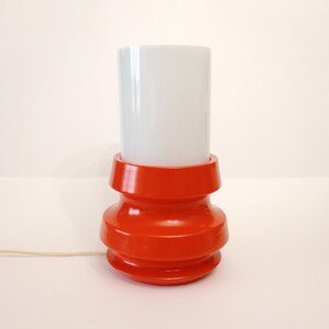Pu&ograve; includere: Una lampada da tavolo rossa con un paralume cilindrico bianco. La base della lampada ha un design unico e a strati.