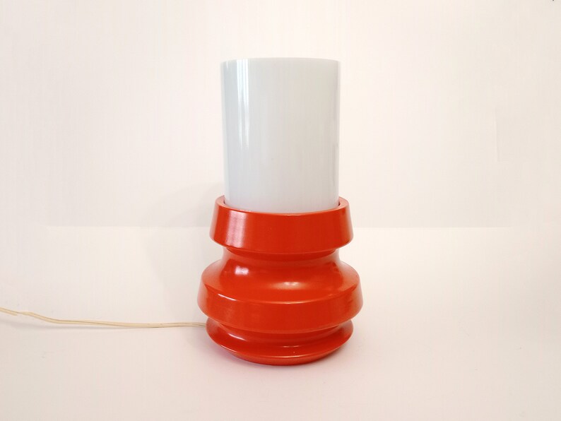 Pu&ograve; includere: Una lampada da tavolo rossa con un paralume cilindrico bianco. La base della lampada ha un design unico e a strati.
