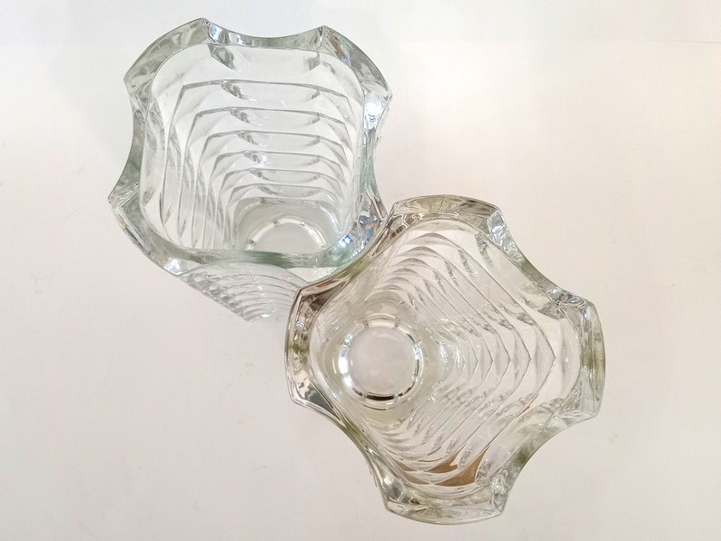 May include: Two clear glass vases with a wavy, textured design. The vases have a square shape with curved edges. The interior of the vases have a series of horizontal ridges. The vases are empty and sit on a white surface.