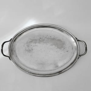 Può includere: Un vassoio da portata ovale color argento con una superficie liscia e riflettente caratterizzata da una patina leggermente ossidata. Il vassoio presenta due manici curvi e decorativi sui lati opposti e un bordo rialzato ben definito.