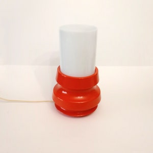 Pu&ograve; includere: Una lampada da tavolo rossa e bianca con un paralume cilindrico bianco e una base rossa con un design a coste.