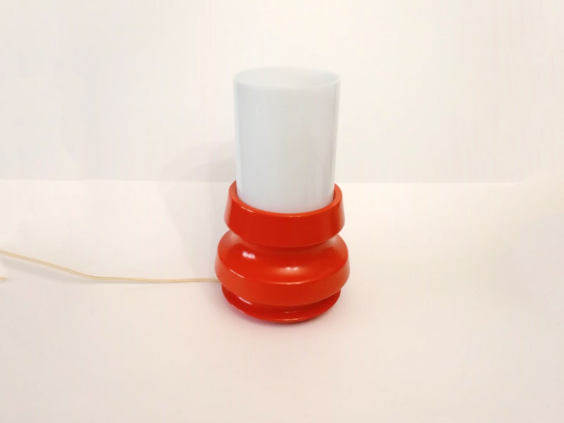 Pu&ograve; includere: Una lampada da tavolo rossa e bianca con un paralume cilindrico bianco e una base rossa con un design a coste.