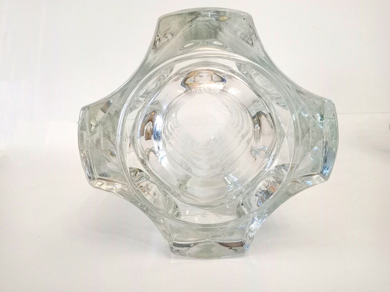 May include: Clear glass vase with a unique geometric design. The vase has a square base that expands outward, with a series of curved edges. The interior is visible, and the word "FRANCE" is etched on the bottom. A decorative piece.