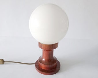 Puede incluir: Una lámpara de mesa moderna de mediados de siglo con una pantalla de vidrio blanco en forma de globo y una base de madera. La base es de color marrón rojizo oscuro y tiene una forma cilíndrica.