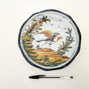 Può includere: Piatto in ceramica dipinto a mano con un disegno di uccello. Il piatto presenta uno sfondo bianco con un'illustrazione dettagliata di un uccello con le ali spiegate, circondato da fogliame giallo e verde. Il piatto ha un bordo blu scuro.