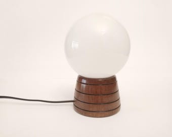 Puede incluir: Una lámpara de globo blanco con una base de madera. La base tiene una veta de madera marrón oscura y está hecha de varios anillos apilados.