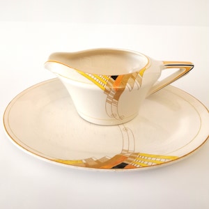 May include: Cream-colored gravy boat and matching plate set. The set features a gold trim and a decorative pattern with yellow, orange, and black accents. The gravy boat has a handle and a spout, and the plate is oval-shaped.