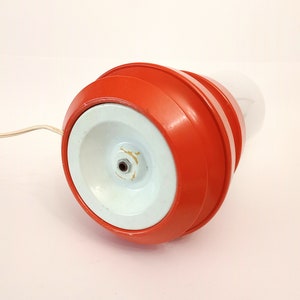 Pu&ograve; includere: Una lampada rossa e bianca con una base rotonda e un paralume bianco. La lampada ha un cavo che si collega a una presa di corrente.