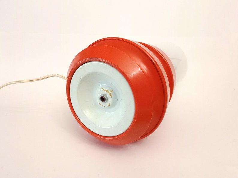 Pu&ograve; includere: Una lampada rossa e bianca con una base rotonda e un paralume bianco. La lampada ha un cavo che si collega a una presa di corrente.