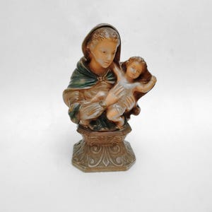 May include: A vintage religious figurine depicting a woman holding a child. The statue features a detailed design with a brown, green, and gold color scheme. The figurine is mounted on a decorative base, showcasing a classic religious theme.