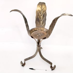 May include: A vintage metal plant stand with a leaf-like design. The stand features a circular base, three curved legs, and a central support with a bowl-shaped holder. The back of the stand has a large leaf shape. The metal has a weathered, aged appearance.