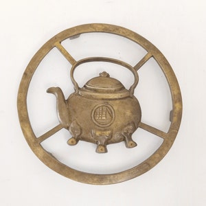 Könnte beinhalten: Ein Teekannen-förmiger Untersetzer aus Messing mit einem runden Design. Der Untersetzer hat ein erhabenes Design einer Teekanne mit Deckel und Henkel. Die Teekanne hat ein chinesisches Schriftzeichen auf der Vorderseite.